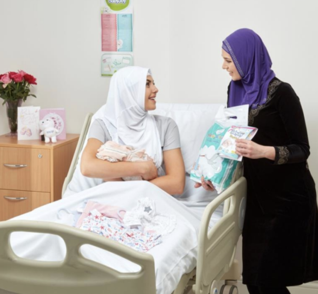 A women approaches another woman in hospital holding her newborn baby. She is showing her different nappy and baby care products.